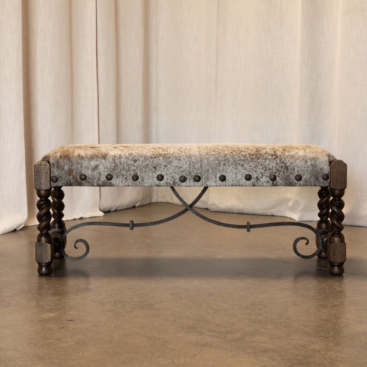 A rustic upholstered bench with a distressed beige and brown velvet seat, dark metal nailhead trim, and twisted dark wood legs on a concrete floor in front of draped cream fabric.