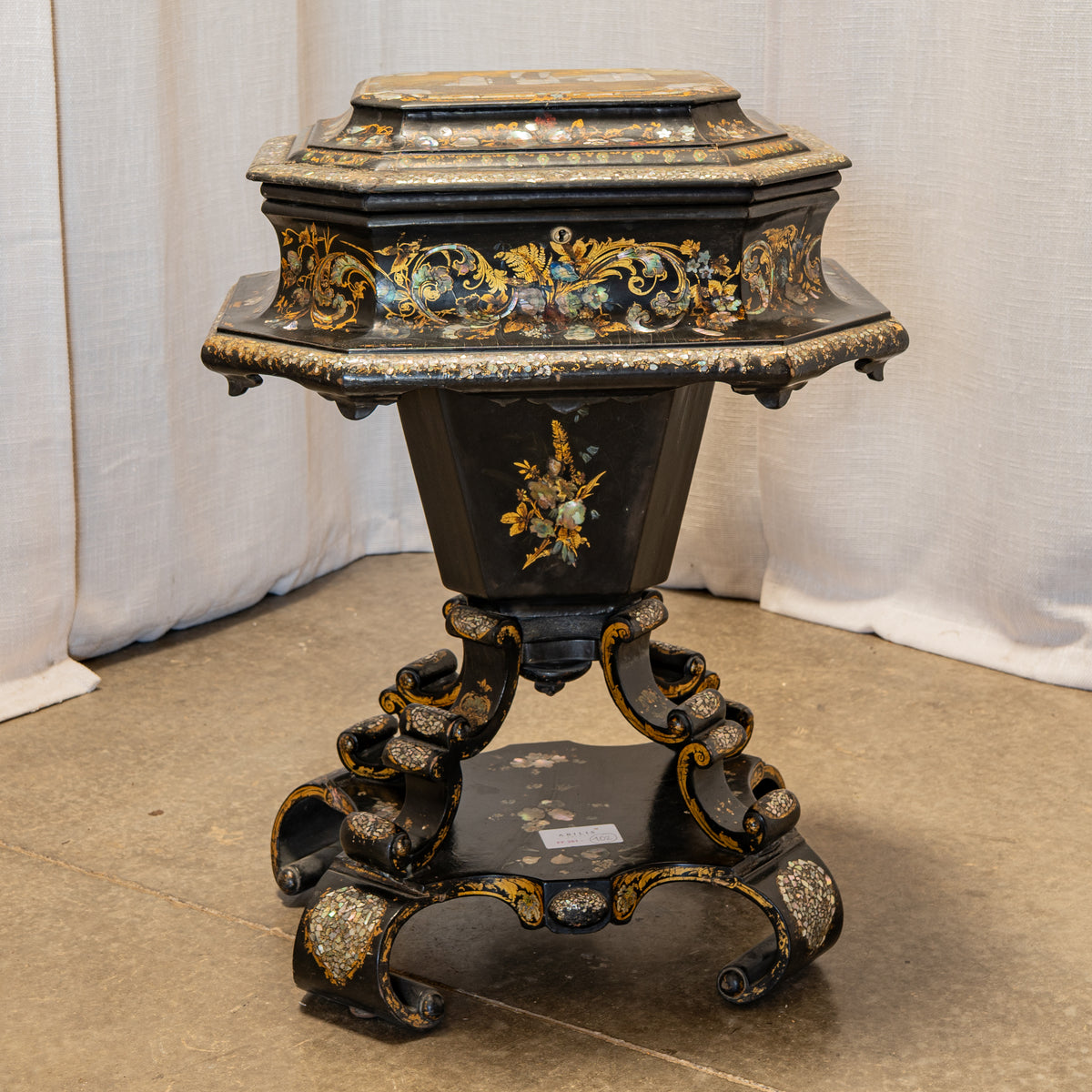 An ornate black lacquered pedestal table with intricate gold and multicolor floral inlay detailing, set against a neutral draped fabric backdrop.