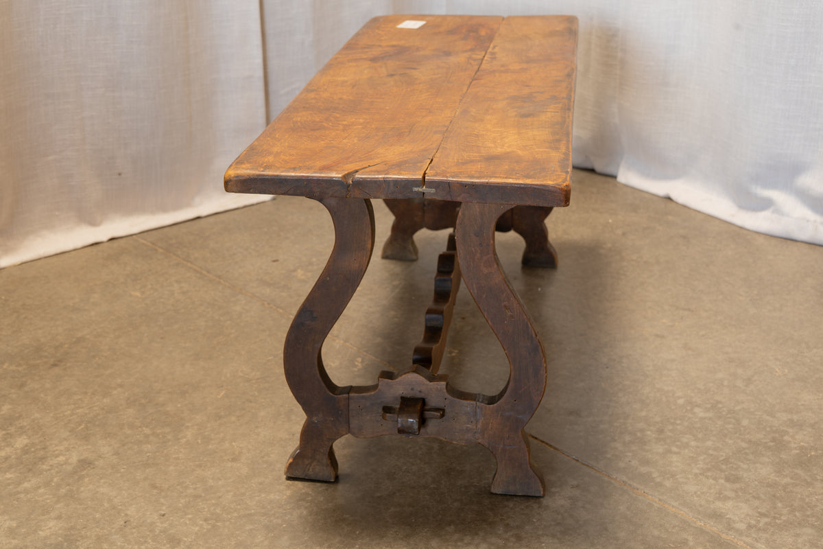 A rustic wooden bench with a weathered brown top and dark carved legs, photographed against a draped white fabric backdrop on a concrete floor.