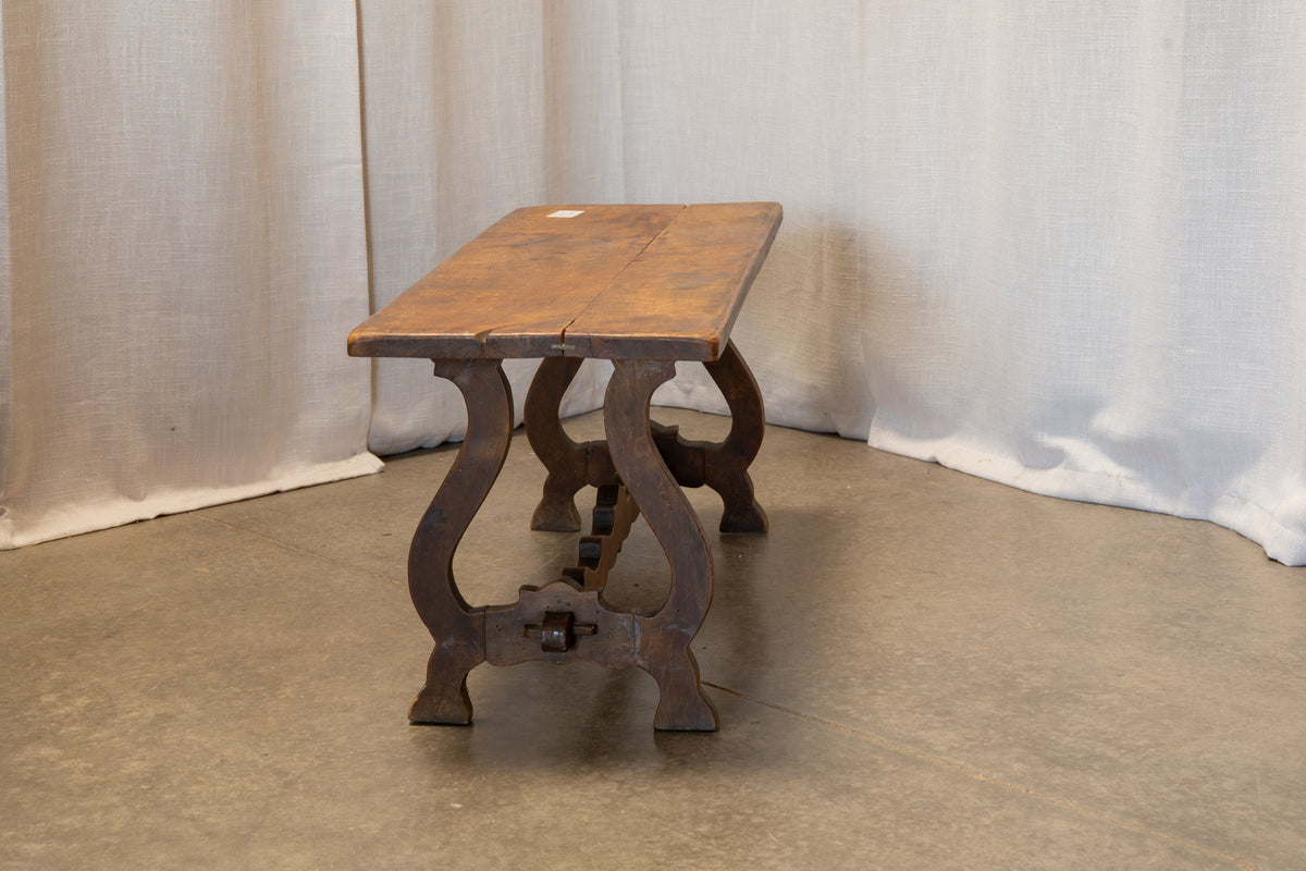 A rustic wooden bench with a rectangular top and curved legs, set against a backdrop of draped white fabric and a concrete floor.