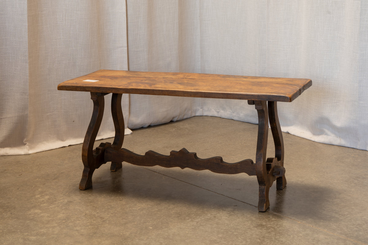 A rustic dark wood console table with a rectangular top, carved legs, and a decorative stretcher bar, set against a neutral draped fabric backdrop.