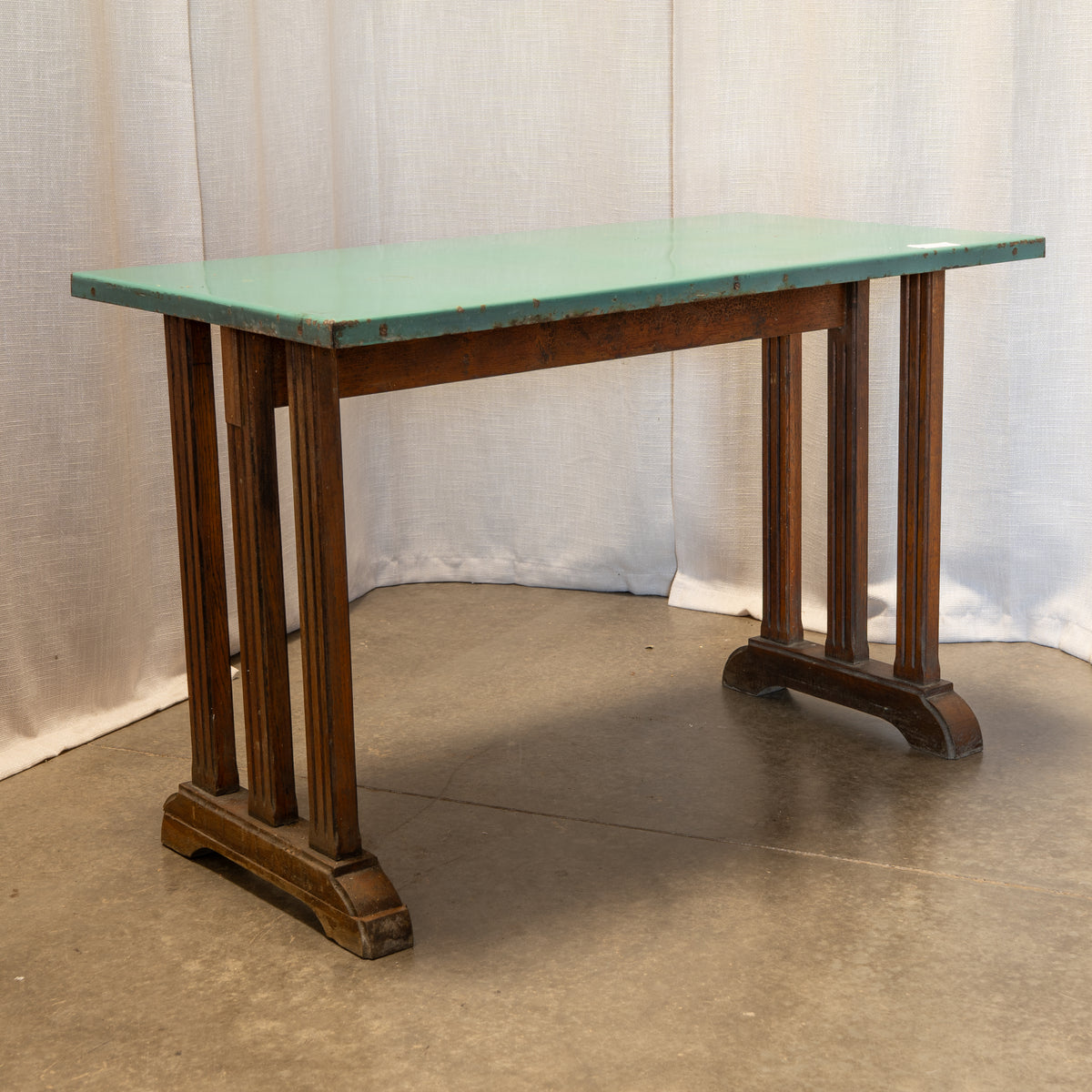 An antique-style writing desk with a distressed mint green top and dark brown wooden legs, set against a draped white fabric backdrop.