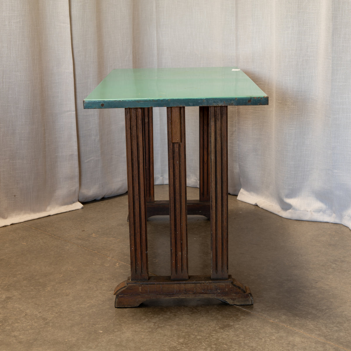 A vintage side table featuring a rectangular mint green enamel top and a dark brown wooden pedestal base with fluted legs, set against a neutral draped fabric backdrop.