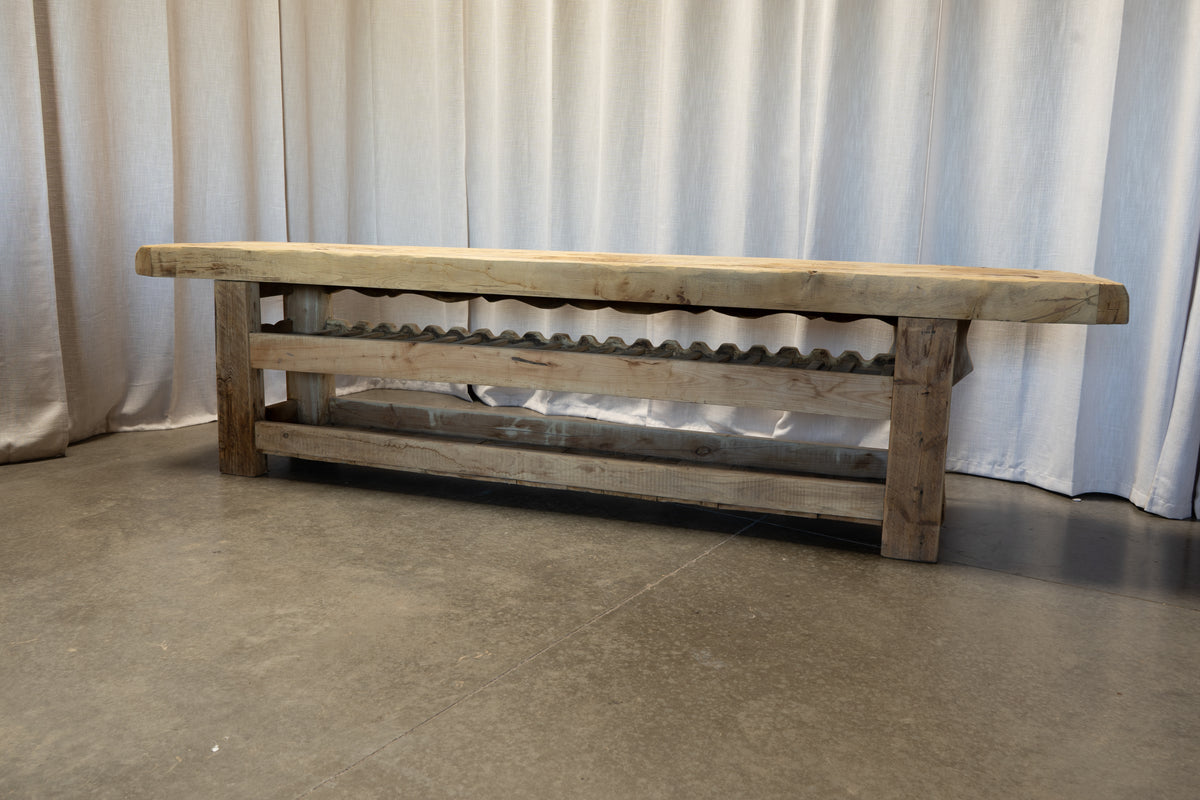 A rustic wooden wine rack bench with a thick natural wood top and weathered gray-brown wooden legs and shelves, set against a white curtain backdrop.
