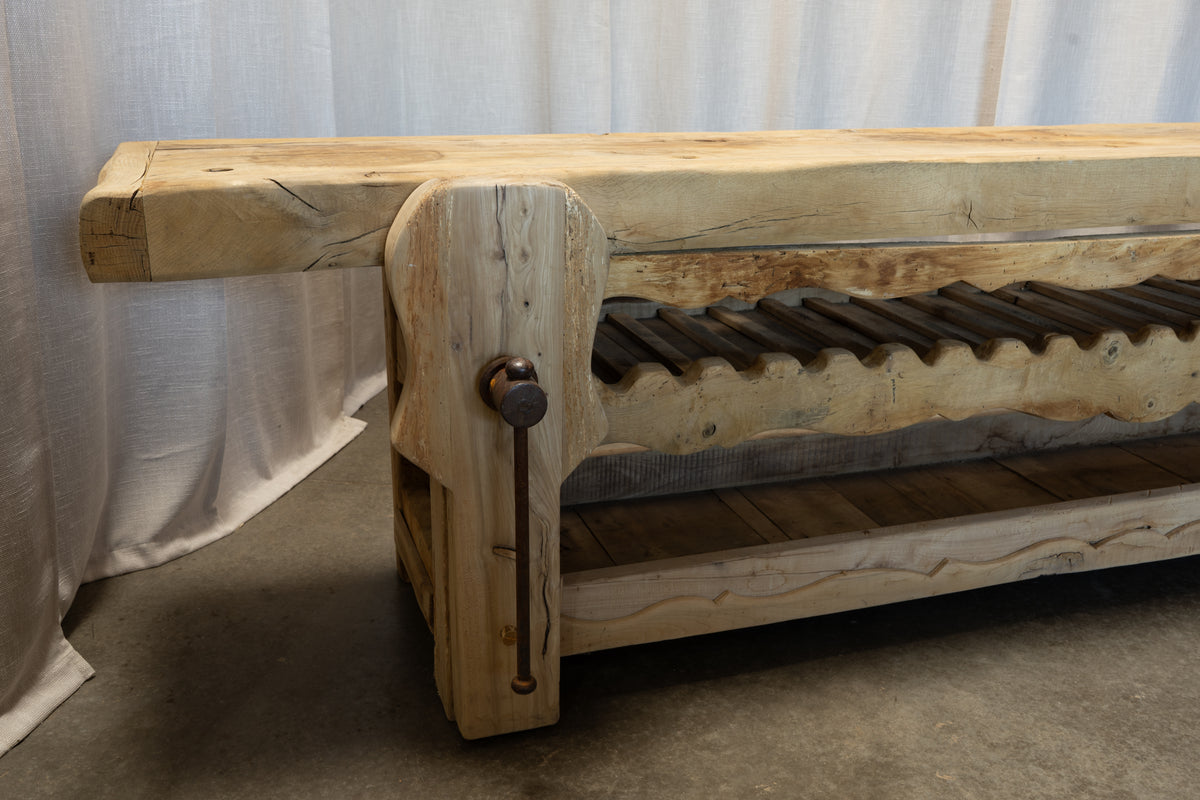 A rustic wooden bench made from a natural live-edge log, featuring a heavy top slab, a crank handle on the front leg, and a slatted lower shelf, set against a draped white fabric backdrop.