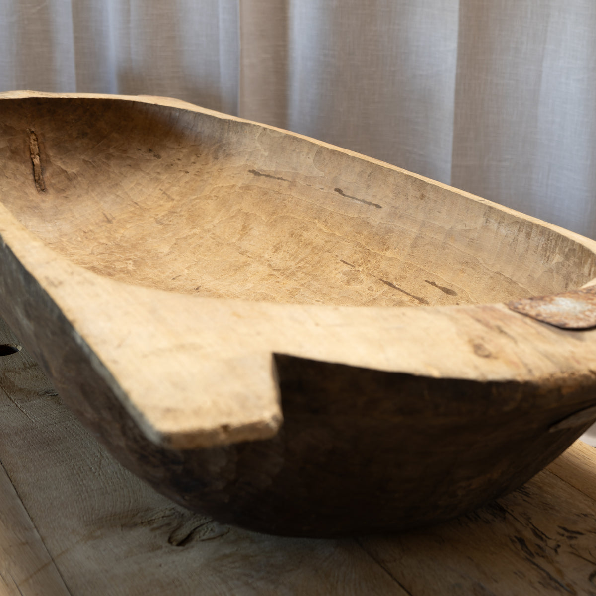 A large, rustic hand-carved wooden trough or bowl with a natural wood grain finish, resting on a light wooden floor in front of neutral beige curtains.