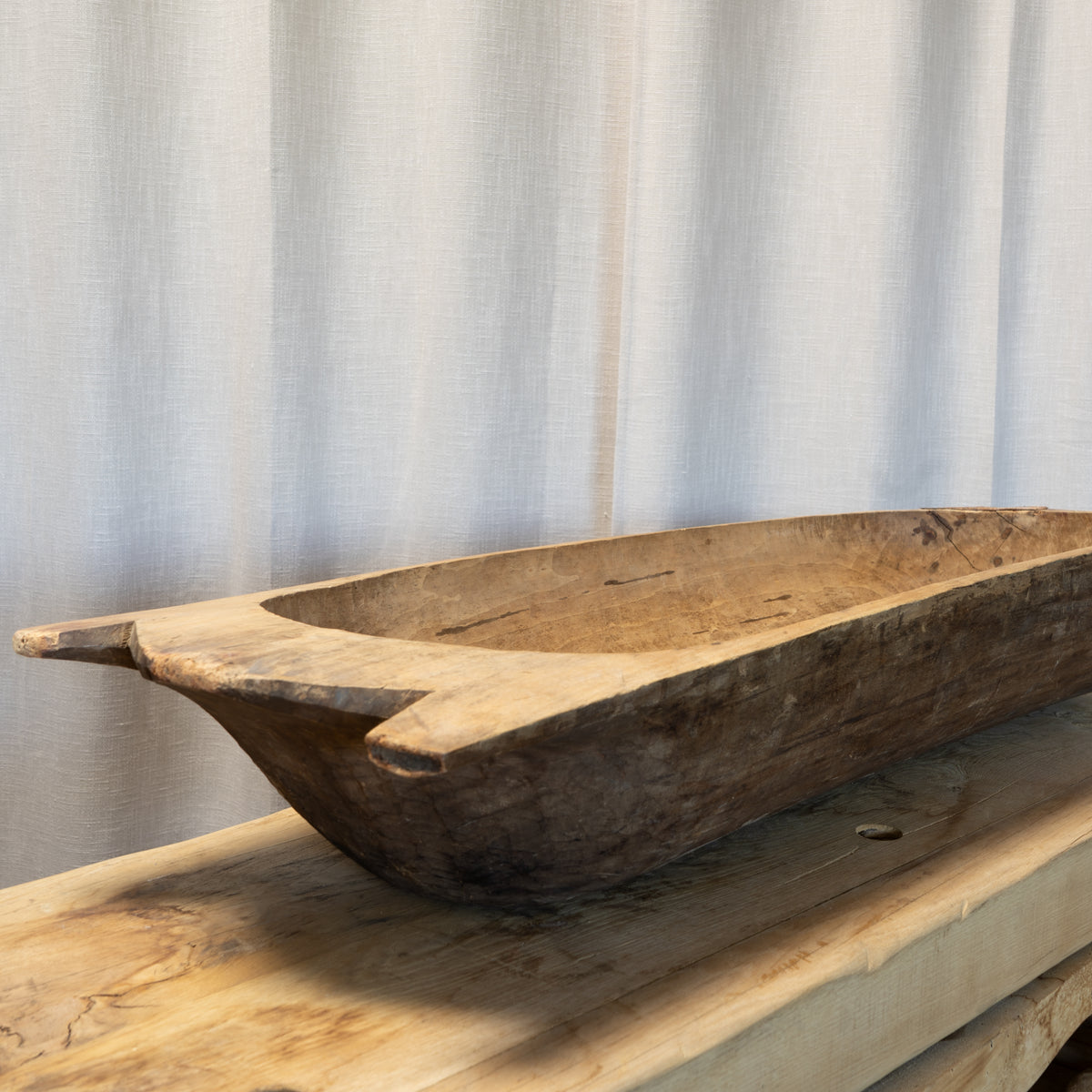 A rustic carved wooden boat-shaped bowl or tray with a natural wood grain finish, displayed on a matching wooden surface against a light beige curtain backdrop.