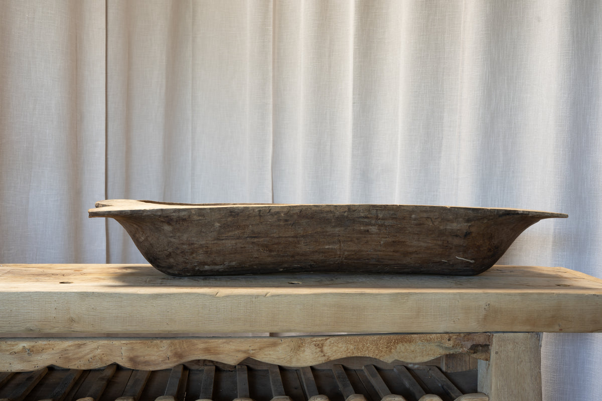 A rustic, hand-carved dark brown wooden trough or planter displayed on a light-colored wooden table against a neutral beige curtain background.