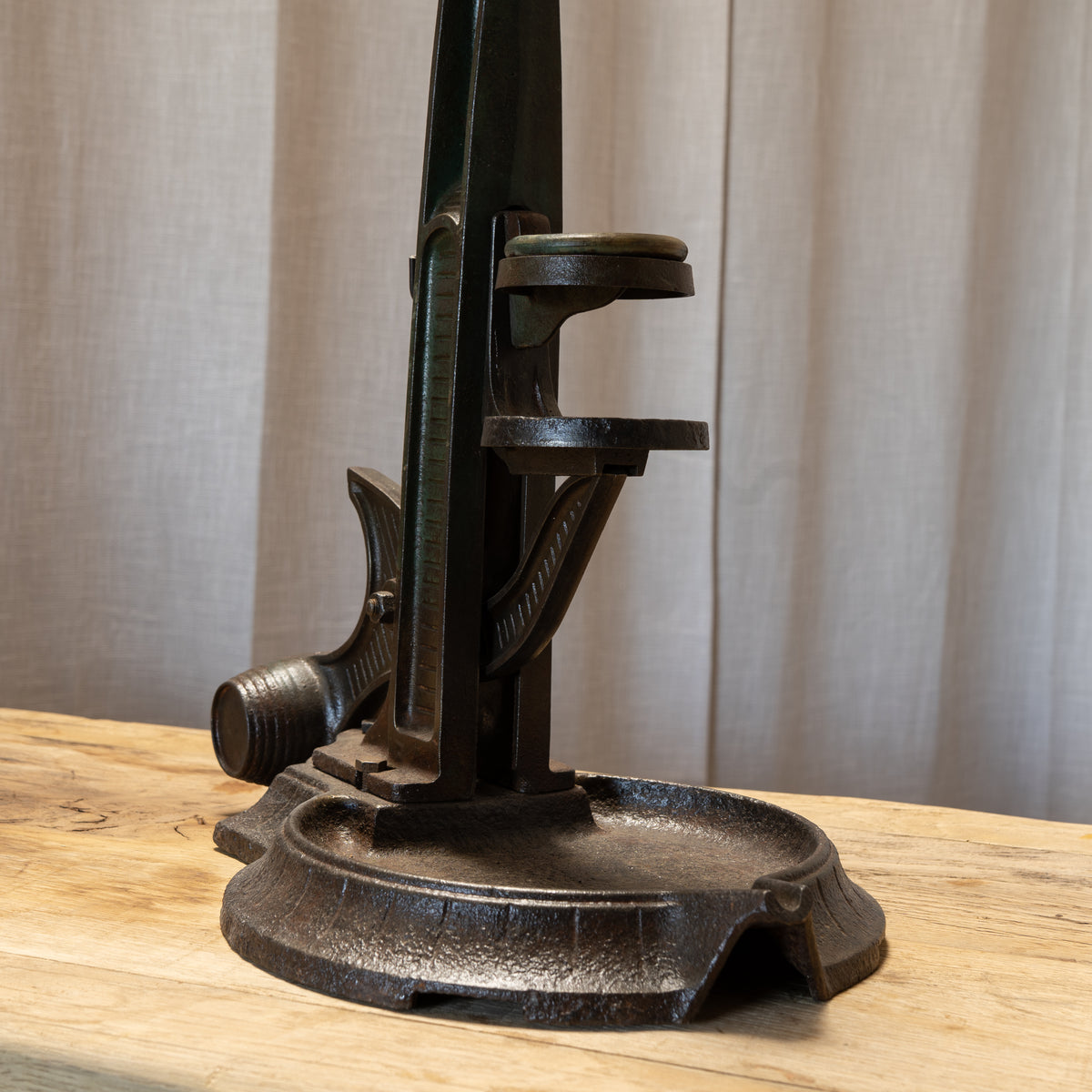 A dark cast iron fireplace poker and ashtray set displayed on a light wood surface in front of a neutral beige curtain backdrop.
