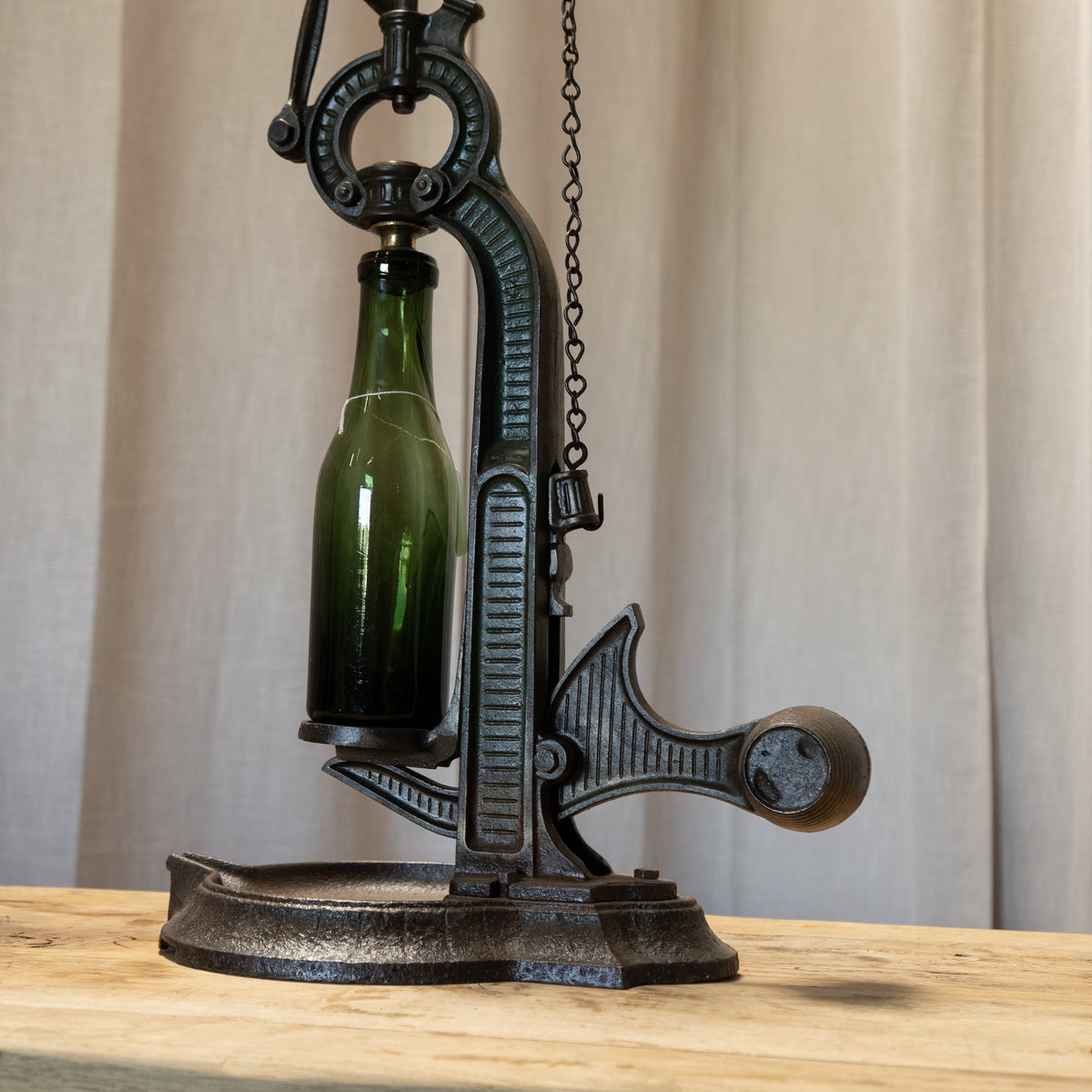 A vintage-style black cast iron bottle opener resting on a light wood table against a neutral beige curtain background. A green glass bottle is inserted into the open top of the iron as a demonstration prop.