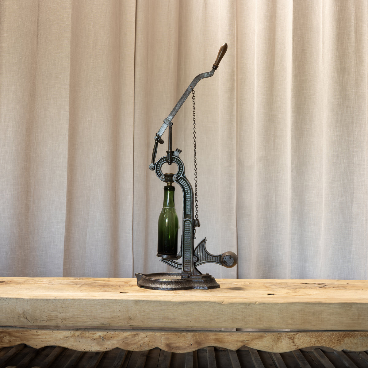 A vintage-style metal bottle opener or press tool with a green glass bottle inserted, displayed on a rustic wooden table in front of beige curtains.