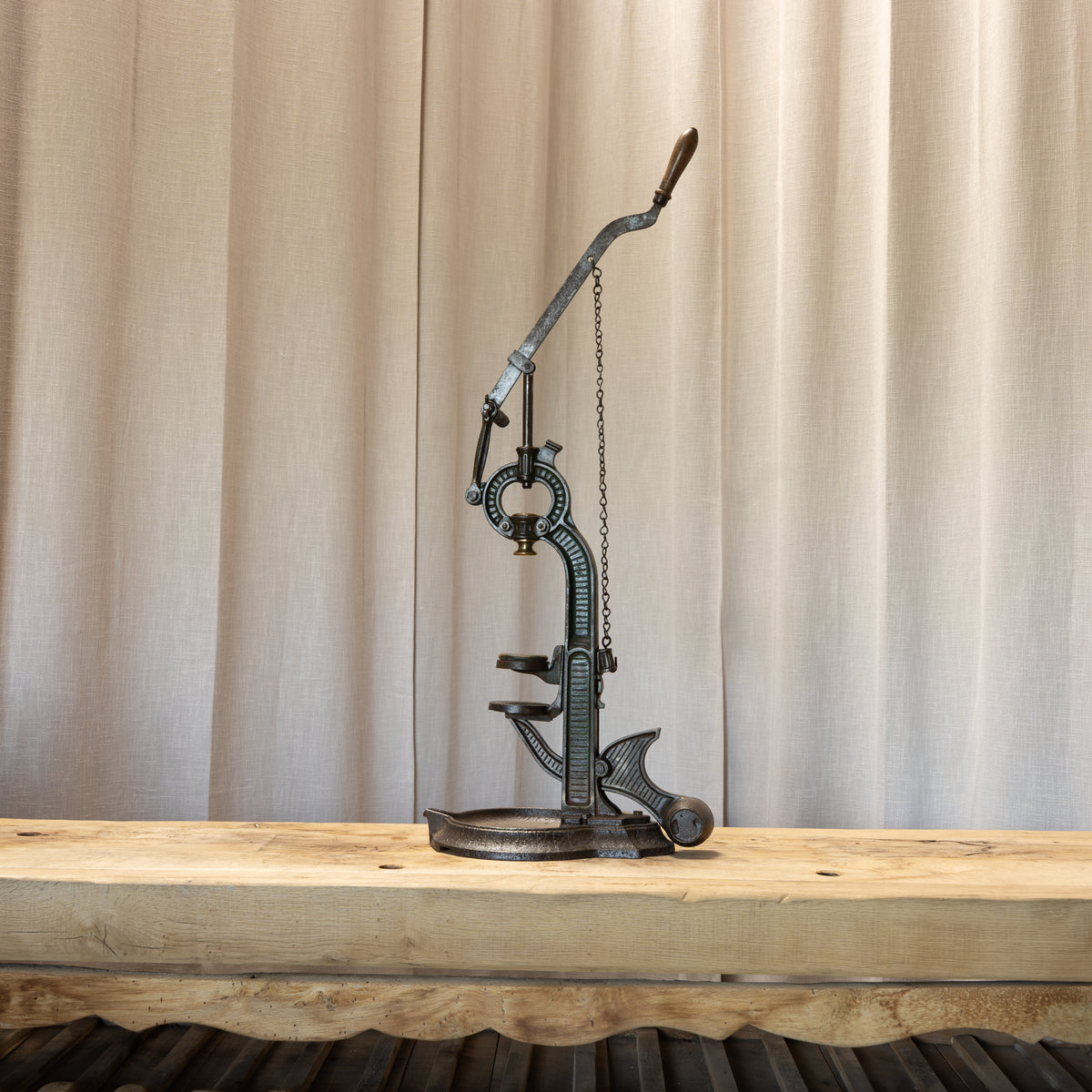 A vintage-style metal press with a long curved handle and chain mechanism, displayed on a rustic wooden table in front of beige curtains.