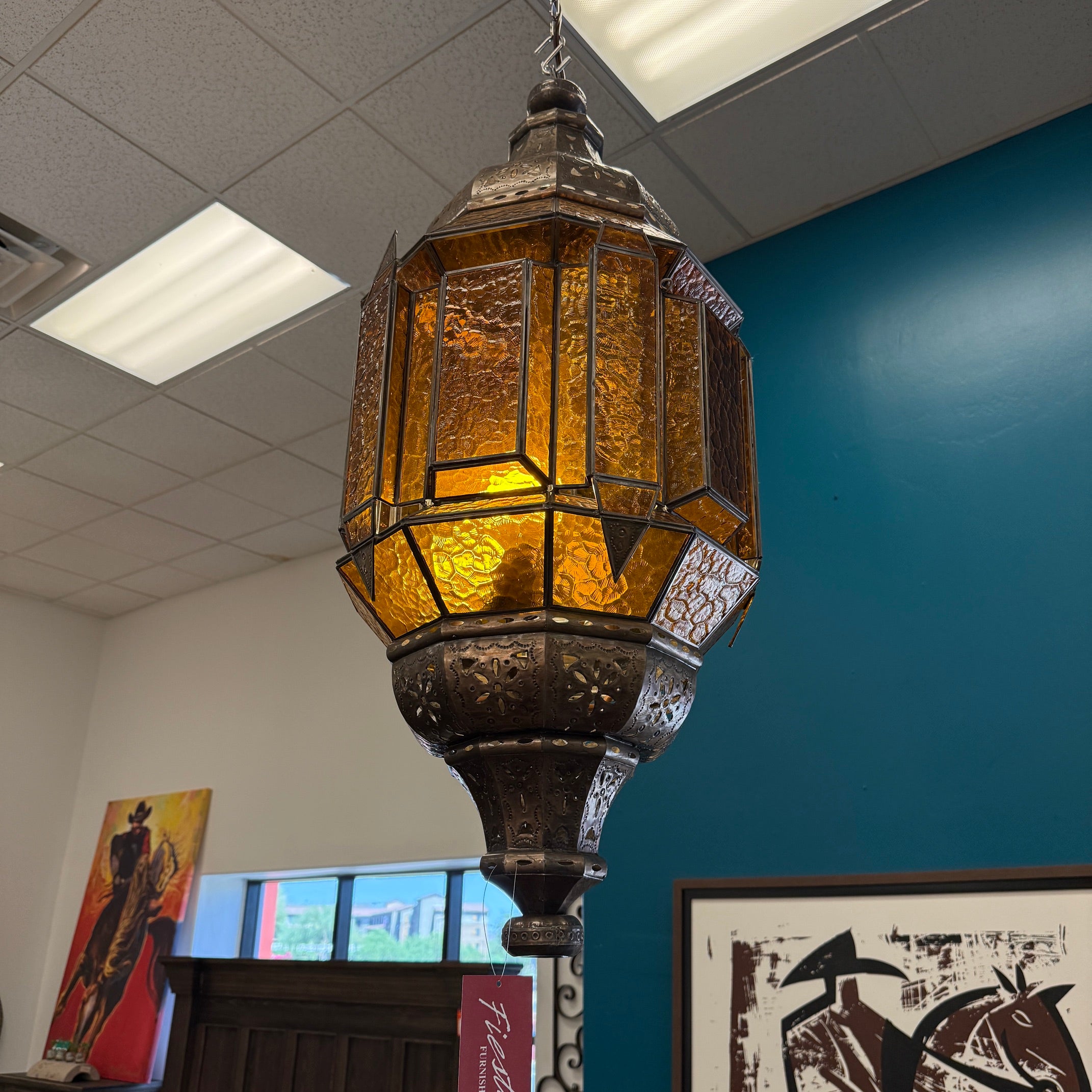 A hanging lantern-style pendant light with a dark metal frame and warm amber-tinted glass panels, shown suspended in an indoor room with a blue wall and ceiling tiles visible.