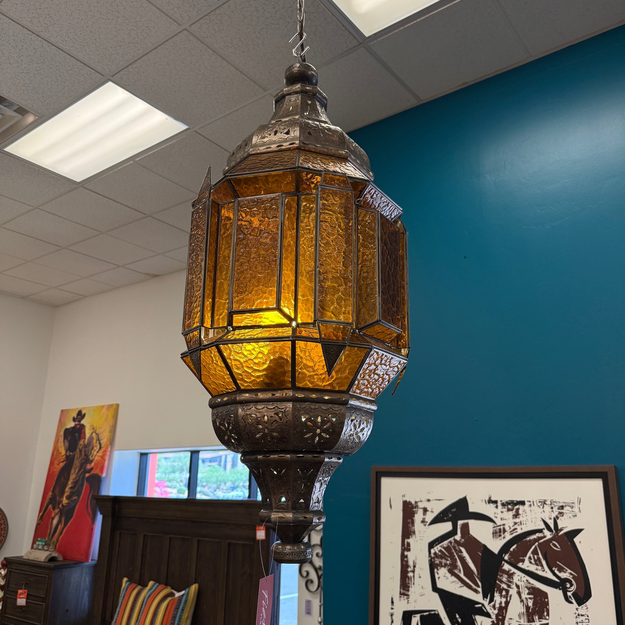 A large hanging lantern-style pendant light with a dark metal frame and amber-tinted glass panels, suspended in a store interior with a blue wall and framed art in the background.