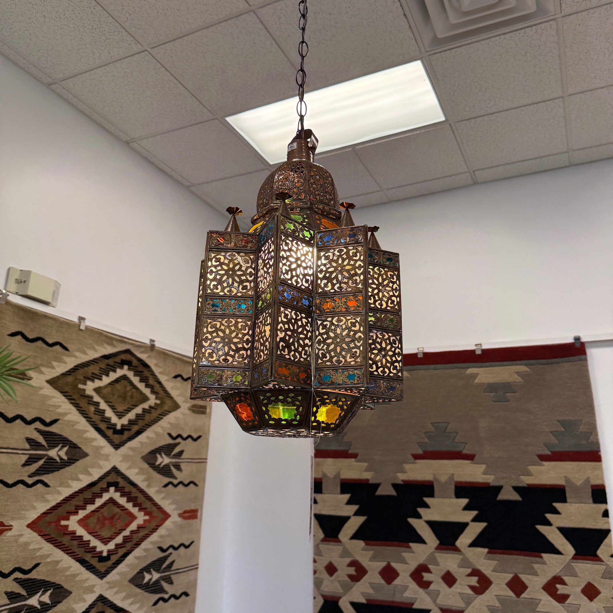 A hanging Moroccan-style metal lantern with intricate cutout patterns and colorful stained glass panels, suspended from a chain in an indoor room with patterned rugs on the wall.