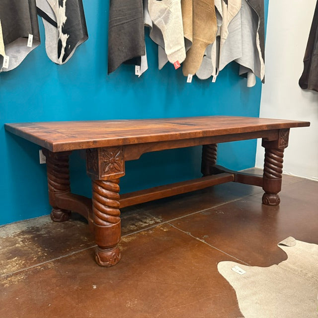 A rustic dark brown wooden dining table with carved twisted legs and a rectangular top, positioned in front of a bright blue wall with hanging textiles.