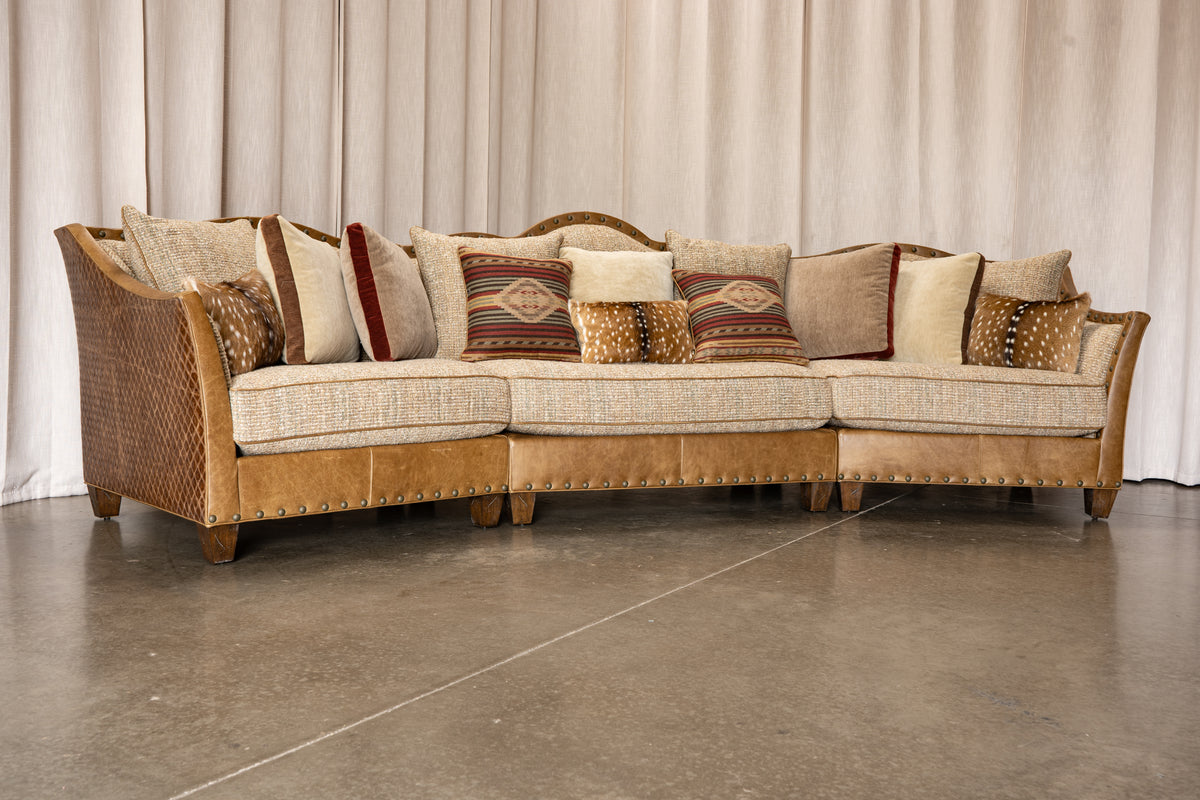 A curved three-seater sectional sofa with woven wicker side panels, beige textured seat cushions, tan leather-like armrests and base, and multiple decorative throw pillows, photographed in a showroom with a beige curtain backdrop and concrete floor.