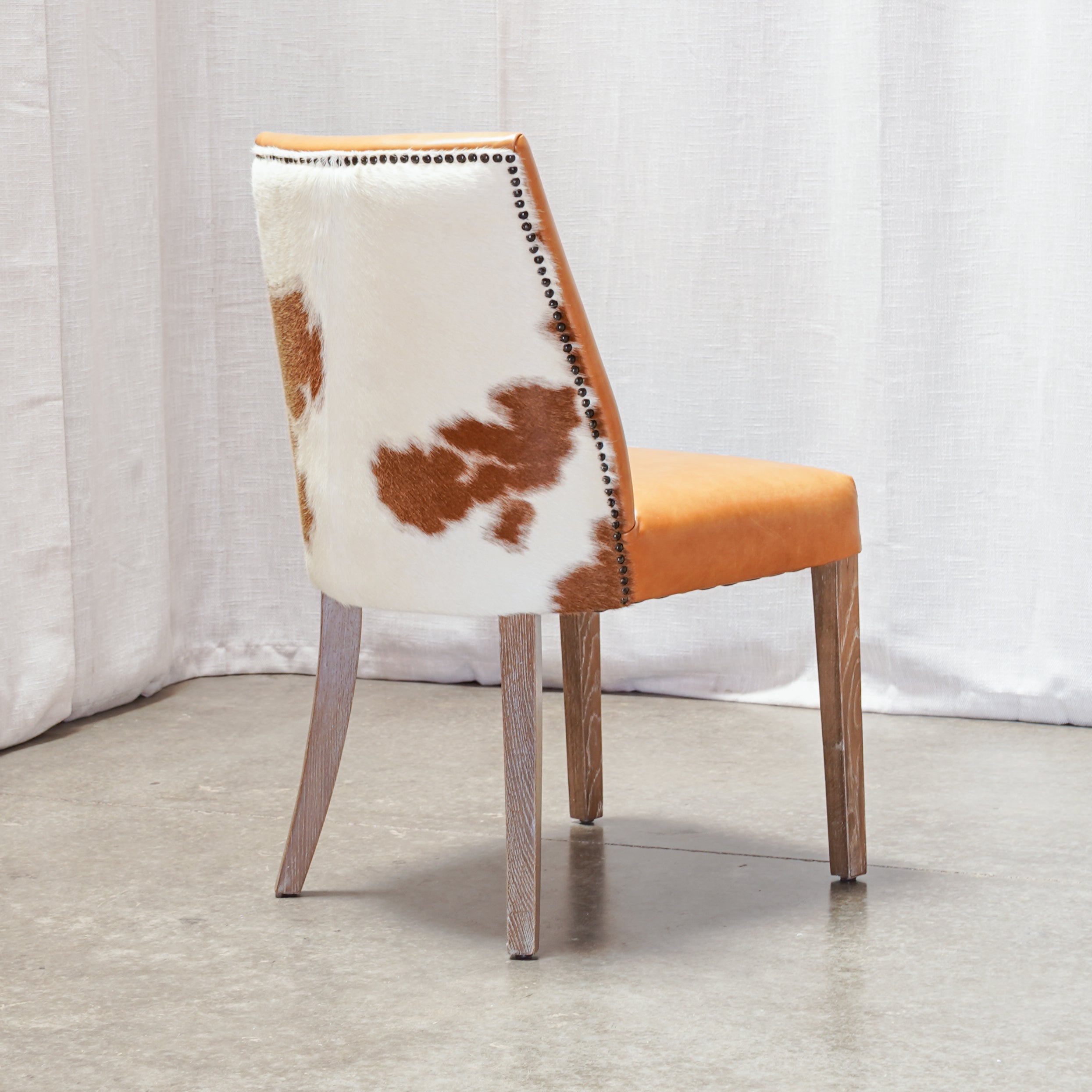 A single dining chair with a cowhide-patterned backrest, orange upholstered seat, and light wood legs, photographed against a white curtain backdrop.