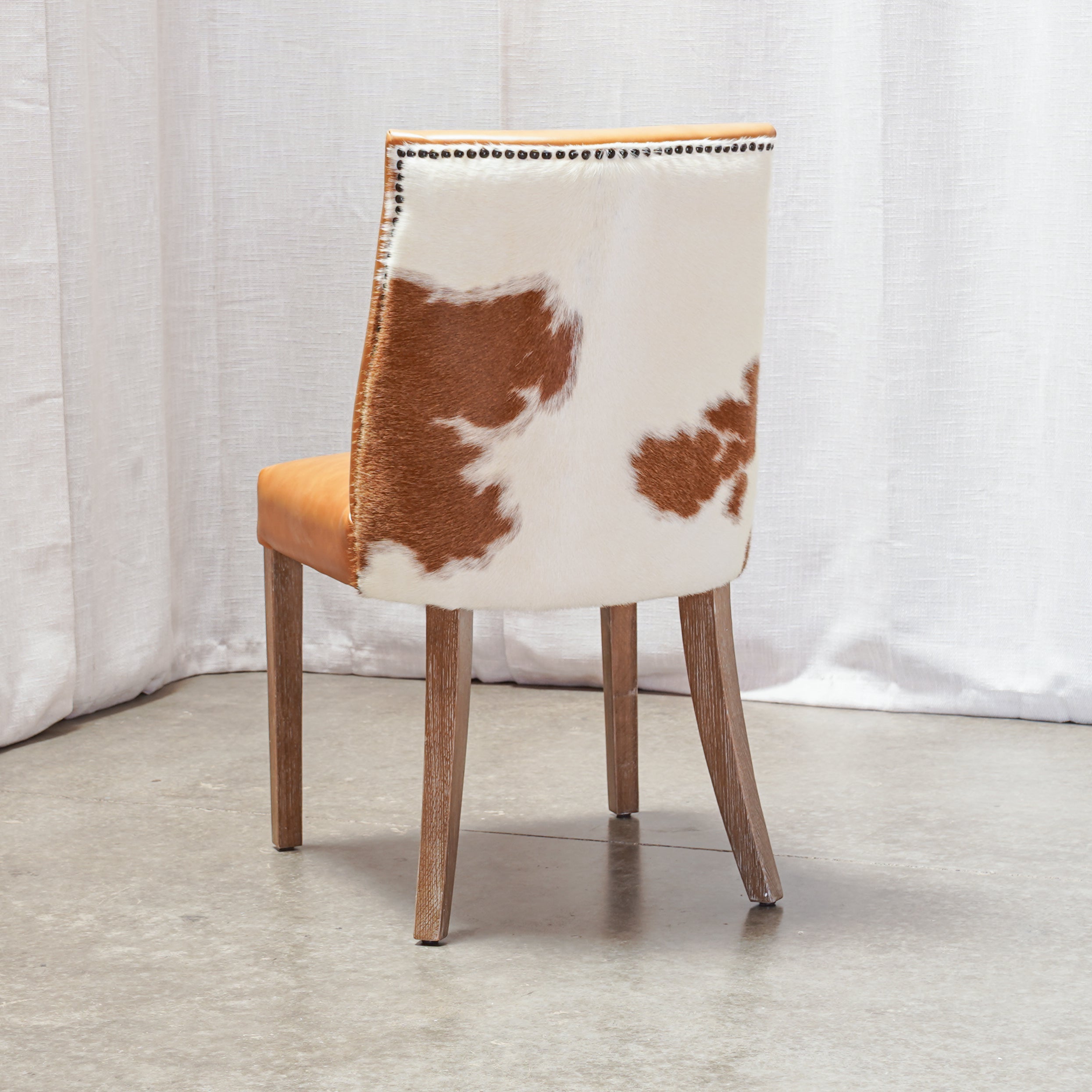 A dining chair with a white and brown cowhide upholstered backrest, a mustard yellow seat cushion, and natural wood legs, shown against a white curtain backdrop.