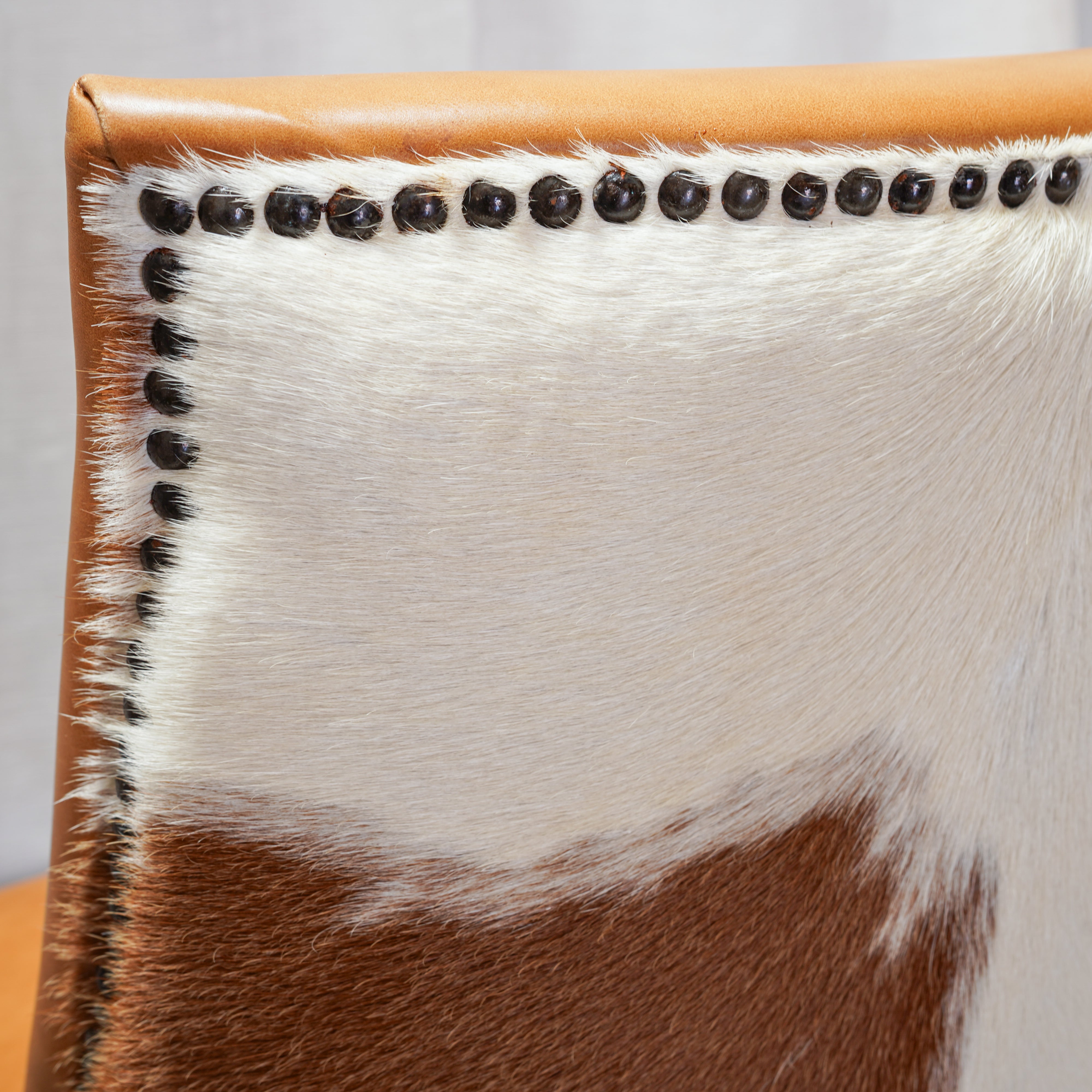 Close-up of a cowhide upholstered chair corner with a tan leather trim, black nailhead trim, and a brown-and-white hide pattern.