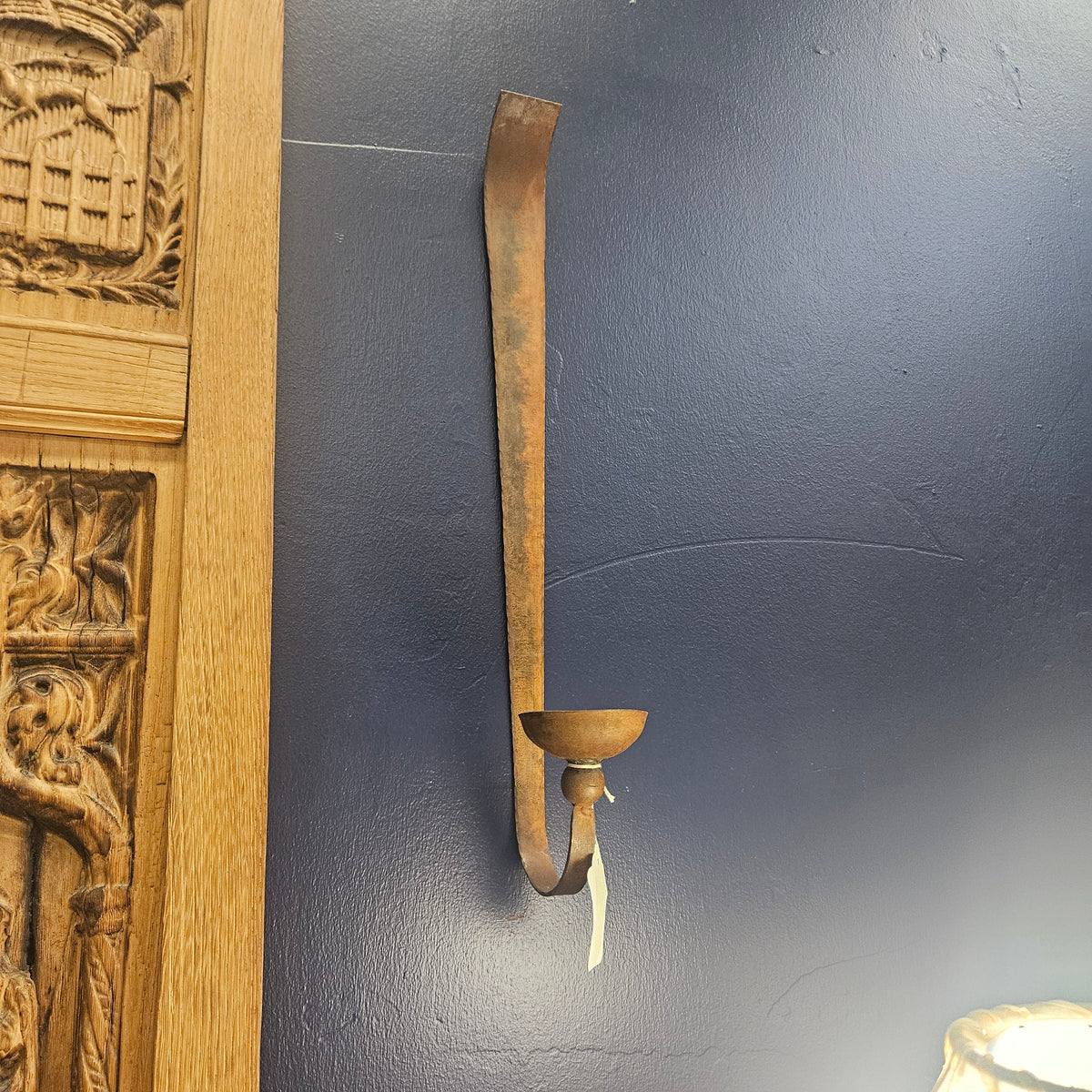 A rustic, curved metal wall sconce with a single candle-style holder, mounted on a dark blue-gray textured wall next to a carved wooden panel.