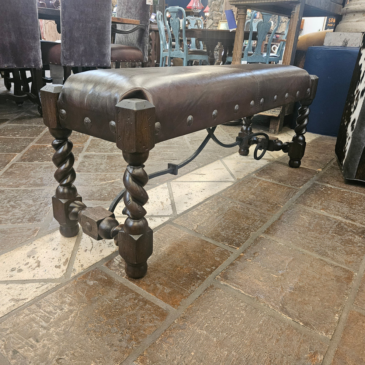A rustic leather bench with dark brown twisted wooden legs and metal studs, photographed in a furniture store on a stone tile floor.