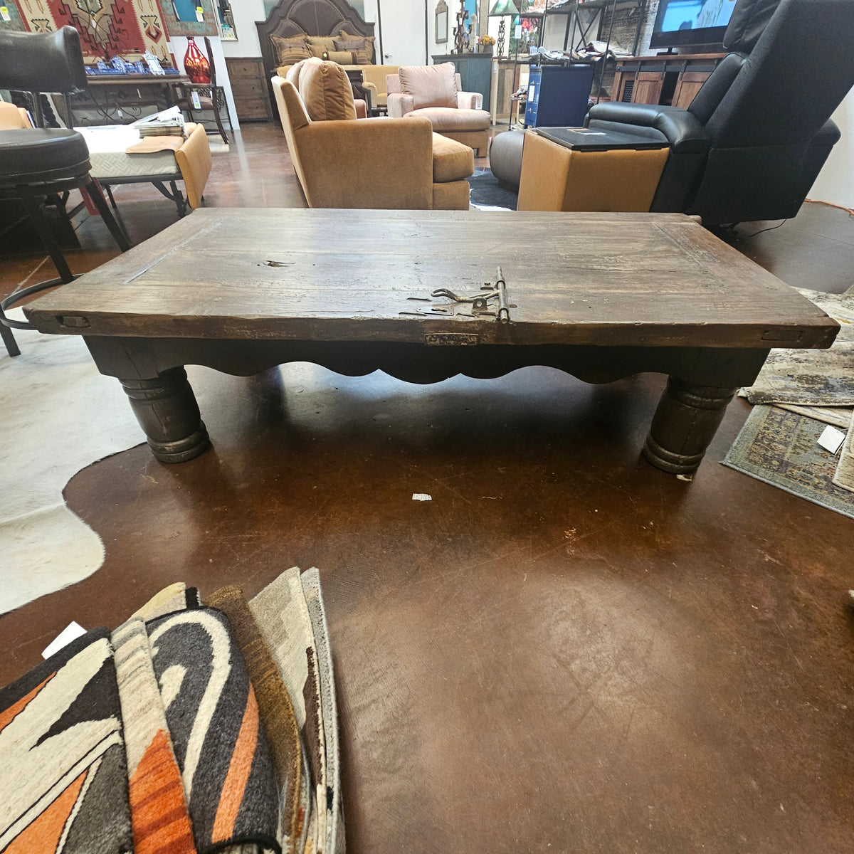 A rustic dark wood coffee table with carved legs and a metal latch on the front, photographed in a furniture showroom with other chairs and decor visible in the background.
