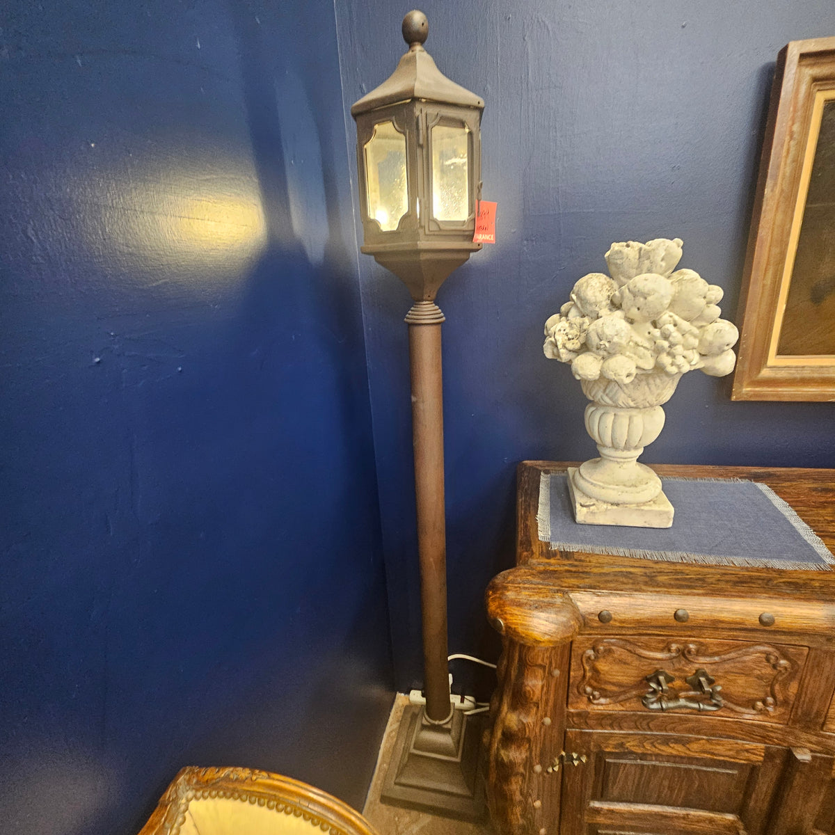 A tall vintage-style floor lamp with a dark metal lantern top and warm glowing light, standing next to a carved wooden cabinet with a white floral urn sculpture on top, set against a dark blue wall.