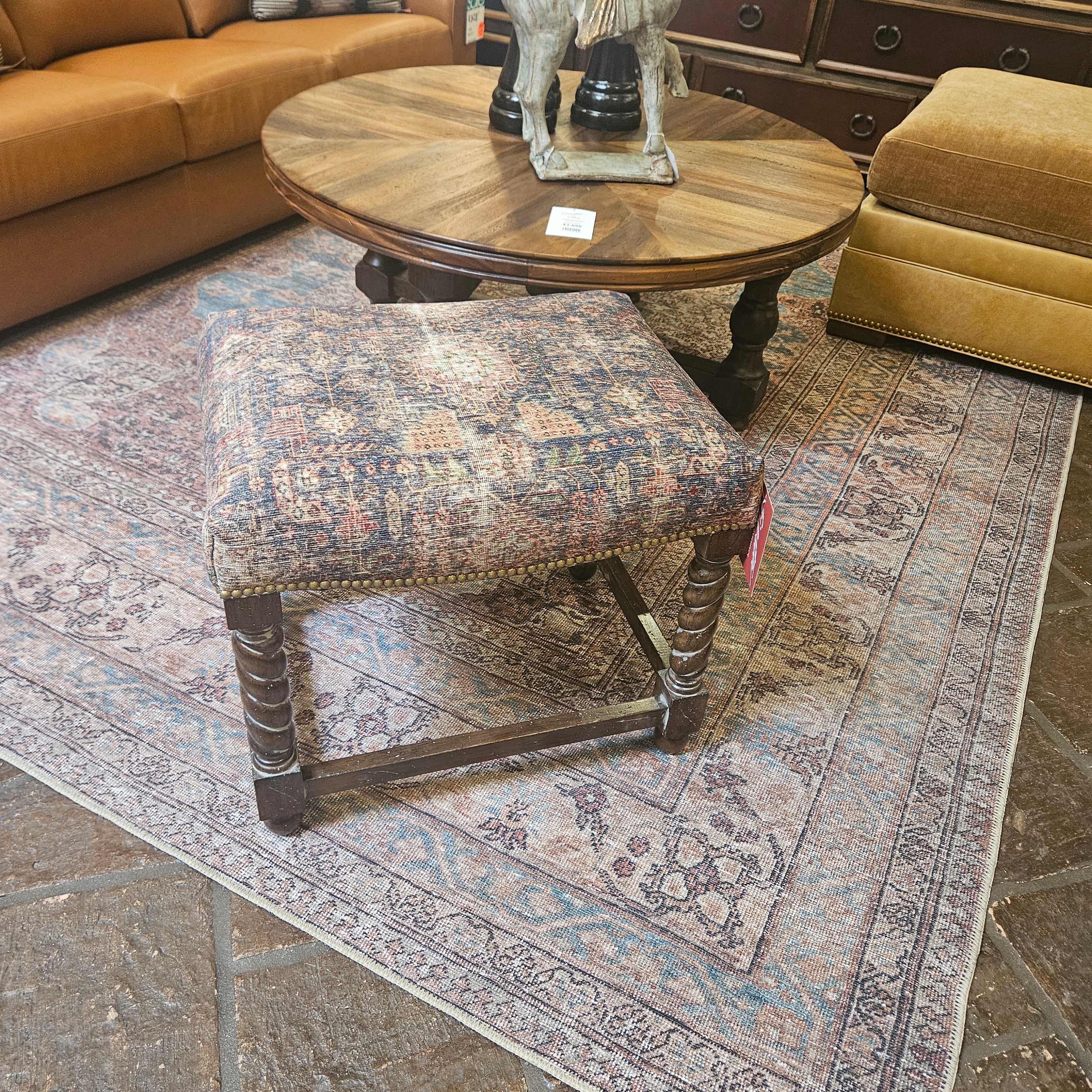 Decorative stool with patterned seat and wooden legs on a textured rug, with a round wooden table and sofa in the background.