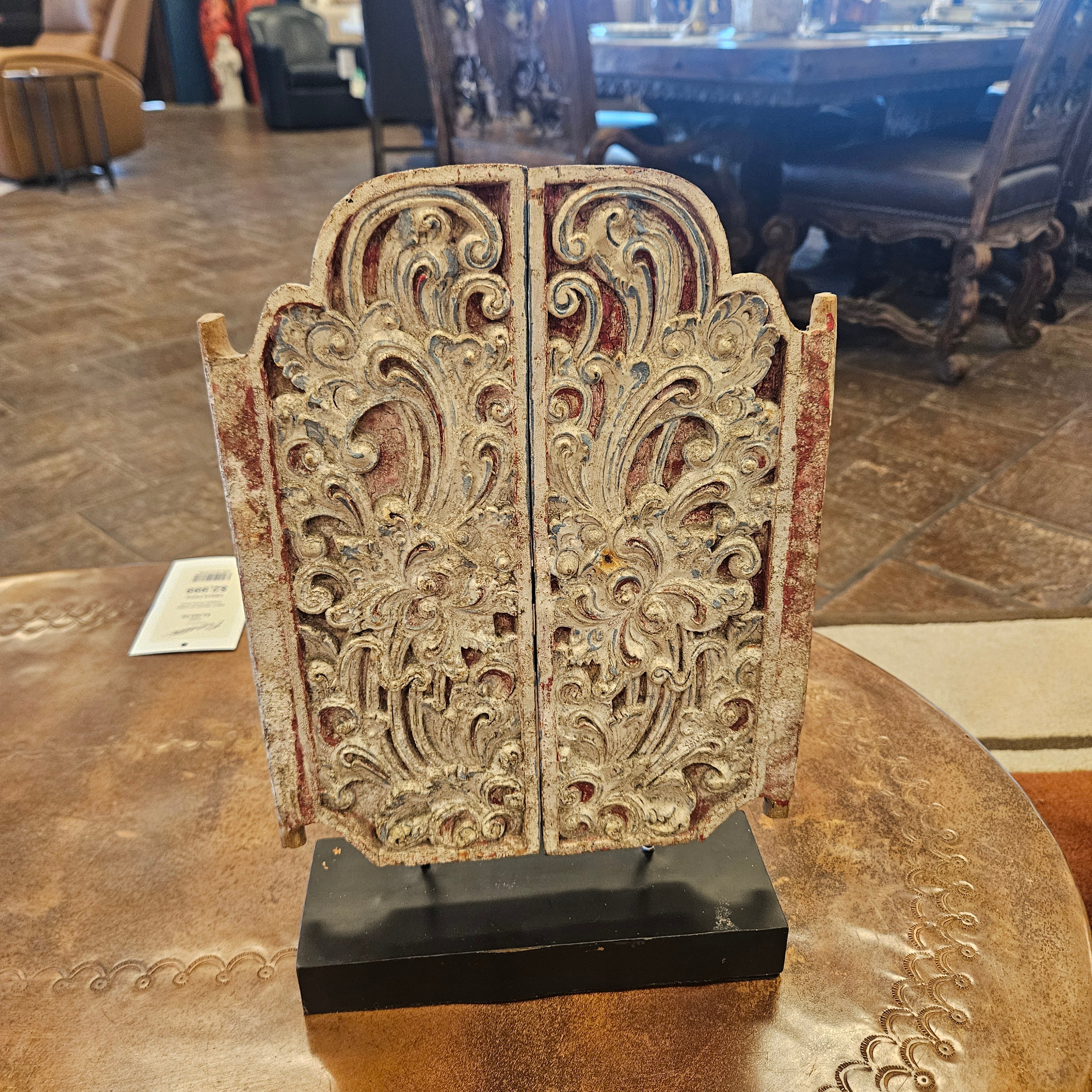 An ornate carved decorative screen or door panel with intricate floral scrollwork, mounted on a black base, displayed on a round wooden table in an indoor showroom.